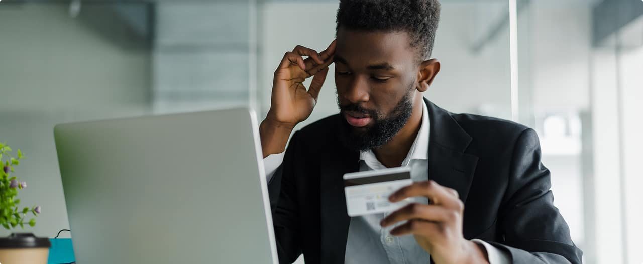 Homem preocupado olhando o notebook enquanto segura cartão bancário após sofrer fraude ou golpe financeiro